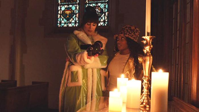 Living for the Dead TV show screenshot 4: Two women stand inside a church holding a video camera. The room is dark side from a cluster of lit candles on a table next to them.