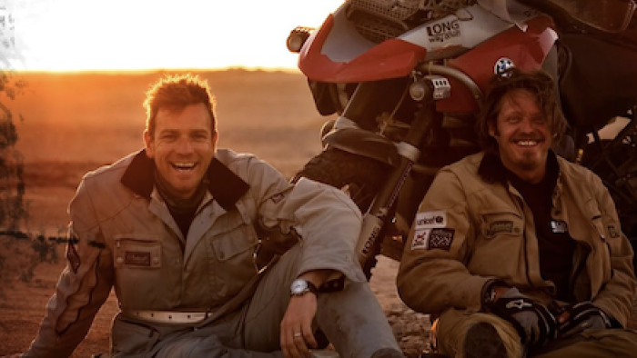 Long Way Down TV show screenshot 1: Ewan McGregor and Charley Boorn sit on ground by motorcycles at sunset.