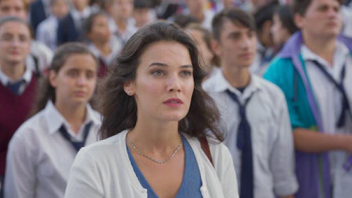 Love 101 TV screenshot 2: Burcu wears a blue top and white cardigan, a group of students behind her in softer focus