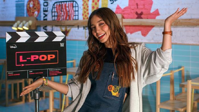 L-Pop TV show screenshot: A young woman is in front of a TV clapper that reads: L-Pop. She stands, smiling with her arms in a "ta-da" pose 