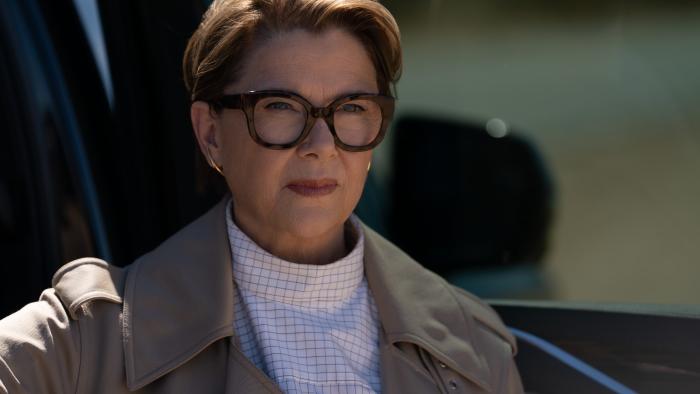 Lucky TV show screenshot 2: Annette Bening stands in an open car door. She wears a striped shirt, a tan coat, and thick, black-rimmed glasses.