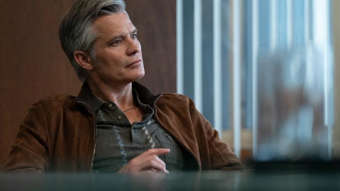 Lucky TV show screenshot 1: Timothy Olyphant sits in a room with dark brown walls. He wears a black suede jacket and a dark gray polo shirt.