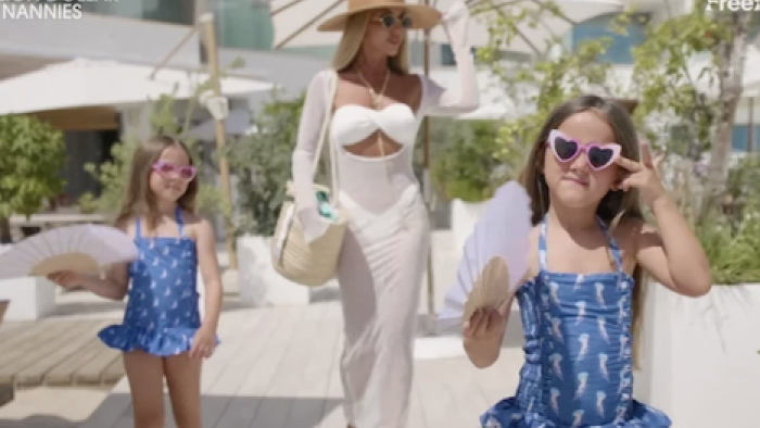 Million Dollar Nannies TV show screenshot 3: Nicola Wills in white dress and hat with two girls in blue swimsuits and sunglasses.
