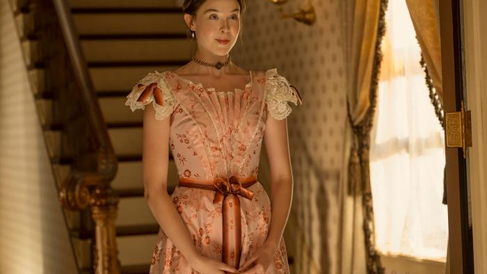 The Lizzie Borden Story screenshot 2: Ella Beatty stands at the bottom of a staircase., wearing a light pink floral dress.