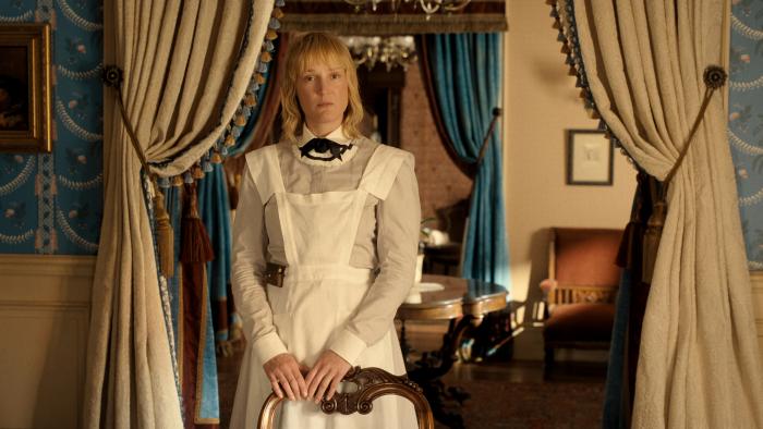 The Lizzie Borden Story screenshot 1: Vicky Krieps wears a light grey dress, with a bow at the collar and apron. She rests her hands on a chair.