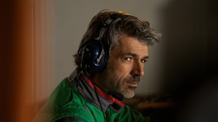 Luca Argentero wears a green, black, and red trimmed pit crew racing jacket. He wears large dark blue over ear headphones.