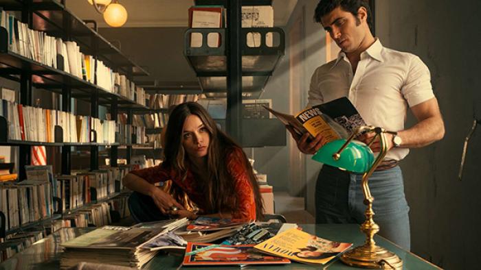 Mrs. Playmen TV show screenshot 1: Elsa and Steve are shown at a desk inside a library, thumbing through news magazines