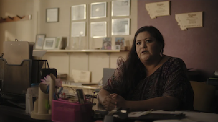 Henny Scott's mother, Paula Castro, a Native woman, covers her mouth with her hand while sitting at a table in her house