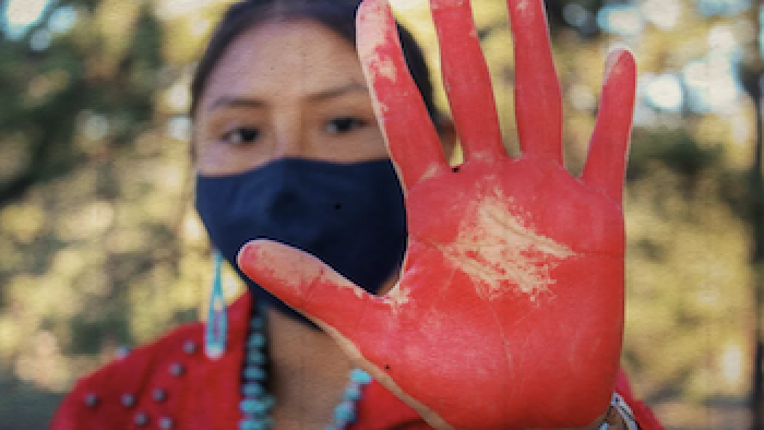 A Native woman wearing red clothing, turquoise jewelry, and a face mask holds a red-painted hand up to the camera
