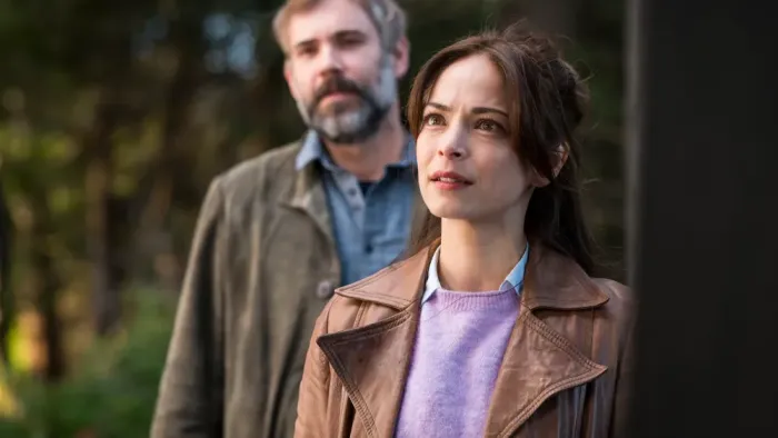Murder in a Small Town TV show screenshot 1: Brunette woman with furrowed brow looks up, listening intently. A man, out of focus, is behind her.
