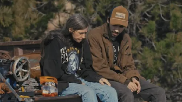 Neighbors TV show screenshot 2: A woman talks to a young man while both sit on the back of a truck. 
