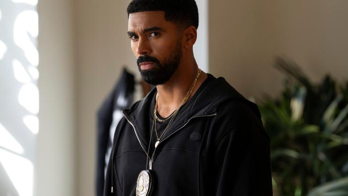 Matthew Law. He wears a black zip up jacket and a black shirt. Around his neck he wears a gold chain, and a police badge.