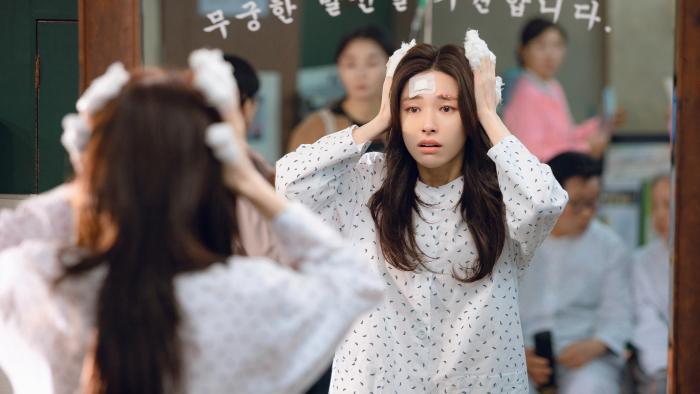 Ha-young holds bandaged fingers against her head as she looks into a mirror. She wears a white hospital gown.