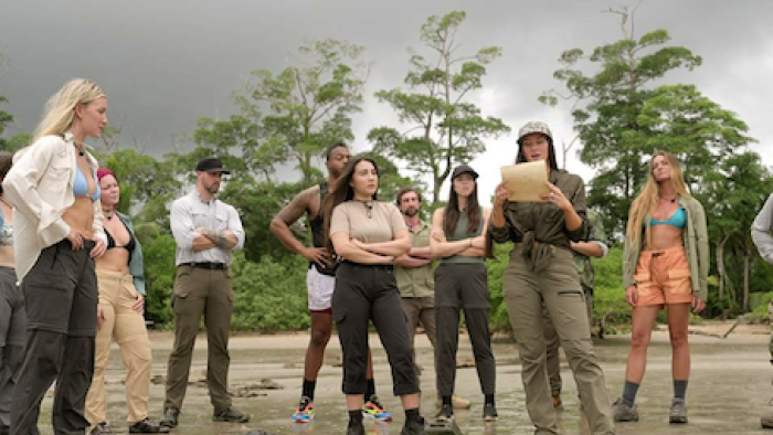 Outlast: The Jungle TV show screenshot 1: Contestants on beach, one reading a paper under a cloudy sky.