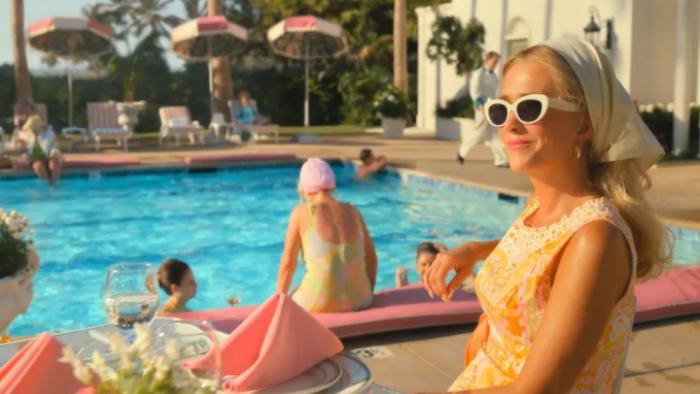 Palm Royale TV show screenshot 1: Maxine smiles by the pool in sunglasses as 1960s styled swimmers are behind her