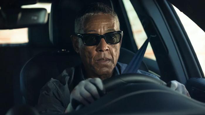 Giancarlo Eposito as Parish sits behind the wheel of a car wearing sunglasses and grey driving gloves