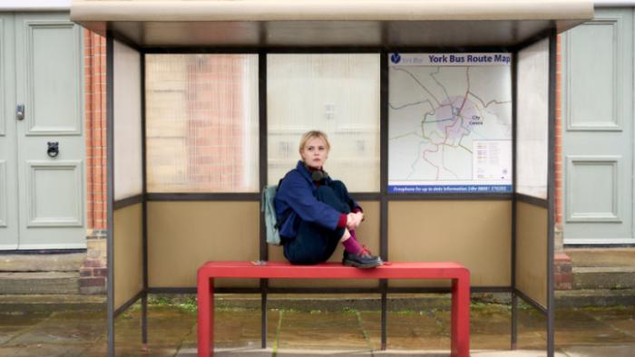 Patience TV show screenshot 3: Patience sits at a bus stop in the daylight with her legs drawn up on the bench