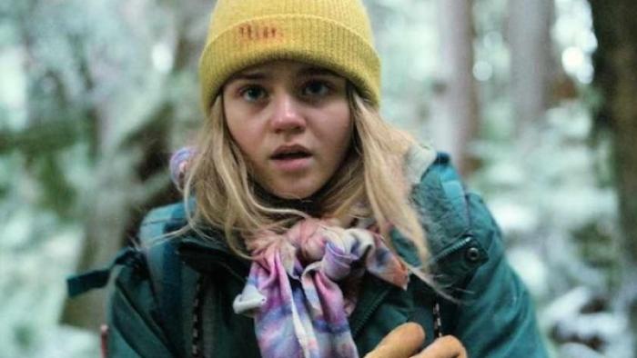 Penelope stands in the woods looking cold, her nose slightly pink. She wears a green coat and yellow hat with blood on it