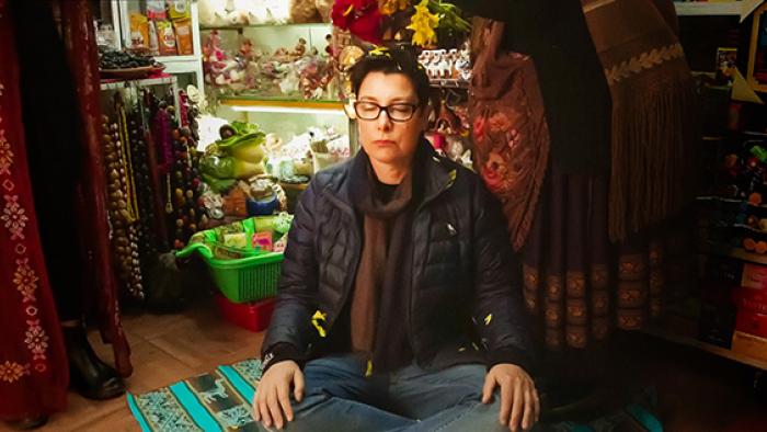 Sue Perkins sits cross-legged on top of a multicolored tapestry, wearing a black puffer coat, with her eyes closed in a meditative pose.