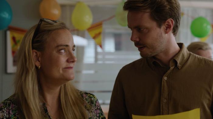 Pernille TV show screenshot 1: A middle aged woman, and slightly younger man look at each other with closed lip smiles in a room full of balloons