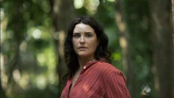 Morgana O'Reilly stands in a forest. She wears a red button-down shirt with thin white stripes.