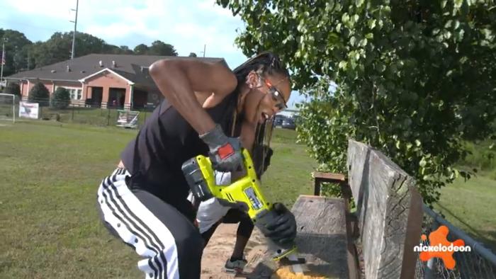 Play It Forward TV show screenshot 2: Renee Montgomery uses a chainsaw to cut through a decaying wood bench, wearing safety gear: glasses and gloves.