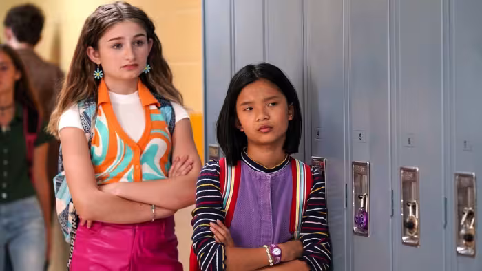Popularity Papers TV show screenshot 1: Lydia (L) and Julie (R) stand serious by the school lockers with their arms folded looking screen-left.