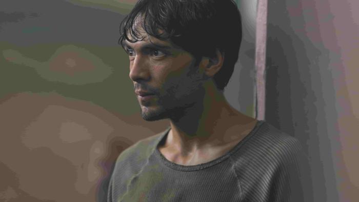 Prison Cell 211 TV show screenshot 1: Diego Calva stands with his back against a pillar. He wears a striped dark grey shirt.