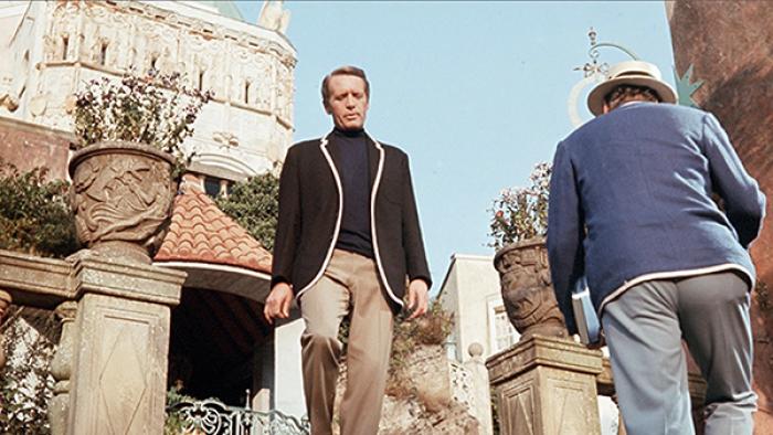 The Prisoner TV show screenshot 1: A man in khaki pants, a black turtleneck, and a black sportcoat with white piping on the lapels is pictured walking down steps outdoors. A man in a similar blue sportcoat and grey pants passes him.