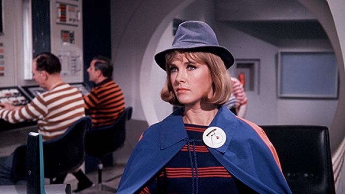 The Prisoner TV show screenshot 4: A woman with a hat and blue cape stands in the foreground. In the background are two men in striped shirts who appear to be working at some kind of computer.