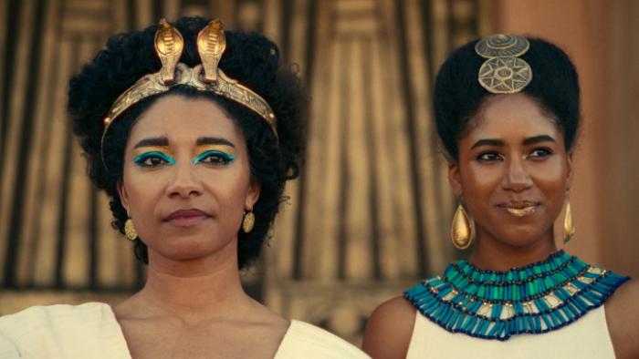 Queen Cleopatra TV show: Cleopatra and Arsinoe are shown onstage during a royal celebration with faces in closeup. Both wear elaborate hairstyles; Arsinoe ha a beaded turquoise collar and Cleopatra has a gold crown with snakes. 