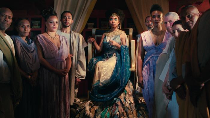 Queen Cleopatra TV show: Cleopatra is in the center of this image on a throne, with advisers and relatives lined up on either side of here, all gazing intently at the camera