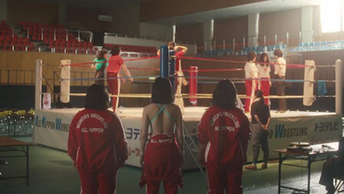 The Queen of Villains screenshot 2: Three aspiring female wrestlers, pictured from behind, stand in a gym watching others work inside the ring