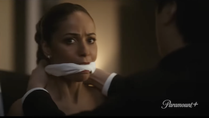 Rabbit Hole TV show: A man with his back to the camera puts a white gag into Meta Golding's mouth