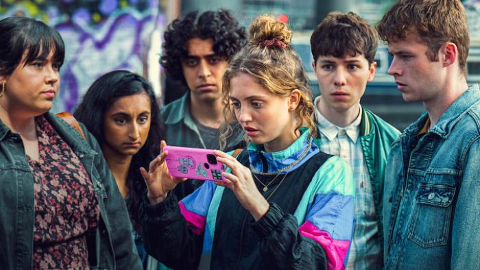 Red Rose TV show: Ash, Jaya, Taz, Wren, Noah, and Antony are grouped together on a street, dressed in street clothing and looking at a phone with absorption