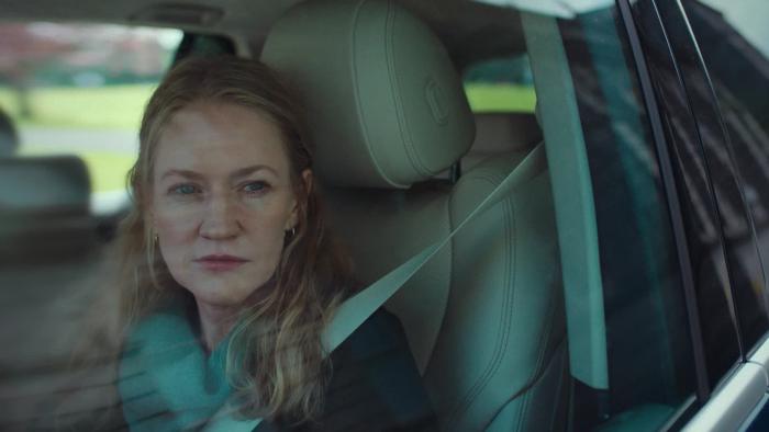 Blonde adult woman buckled into the front seat of a car looking out the window with a smirk on her face.
