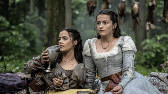  Lydia Peckham and Lauren McQueen sit on blankets. Dead rabbits hang in the background. Peckham holds a chalice near her mouth.