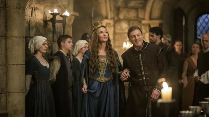 Connie Nielsen and Sean Bean walk in a crowded hall. Nielsen wears a blue dress and a gold crown; Bean wears dark robes.