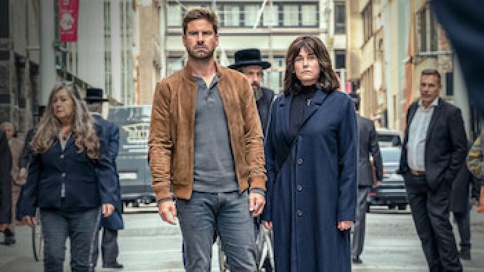 Still from Rough Diamonds: A White man in a jacket and jeans and a White woman in a long coat stand together on a city street.