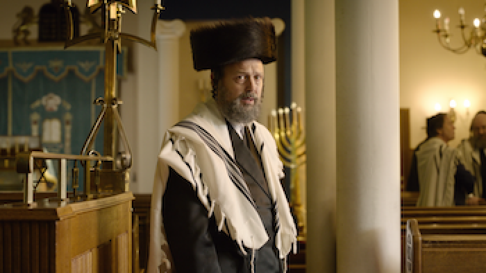 Still from Rough Diamonds: An Orthodox Jewish man stands in a temple with a menorah and other men in the background. 