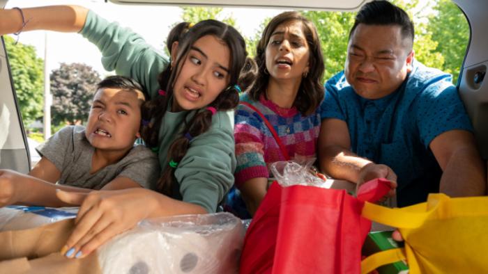 Run the Burbs TV show screenshot 2: All four of the Phams are looking into a car's trunk stuffed with groceries. Leo grimaces with effort; Khia and Andrew are both attempting to pick up heavy bags. 