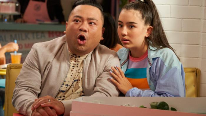 Run the Burbs TV show screenshot 3: Khia has her arm around Andrew as the two sit together at a dining room table. Andrew's mouth is open, seemingly in shock. 