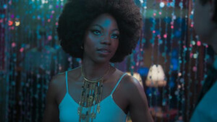Paola González wears a white tank top and an intricate gold necklace. She stands in front of a beaded curtain.