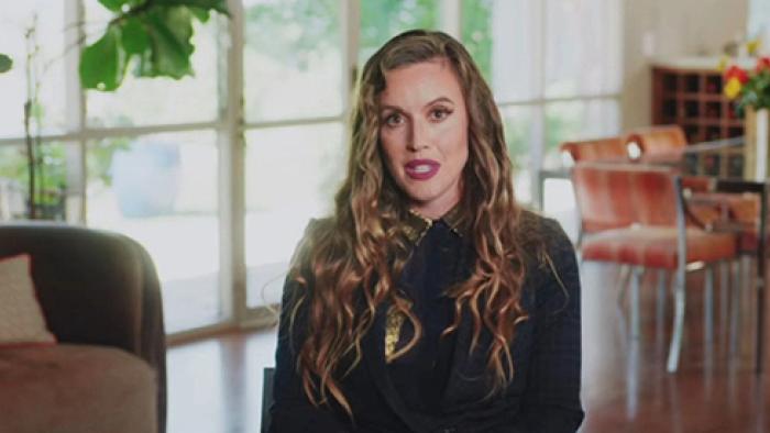 Scamanda TV show screenshot 2: Podcast producer Charlie Webster sits in a chair wearing a dark jacket and deep pink lipstick