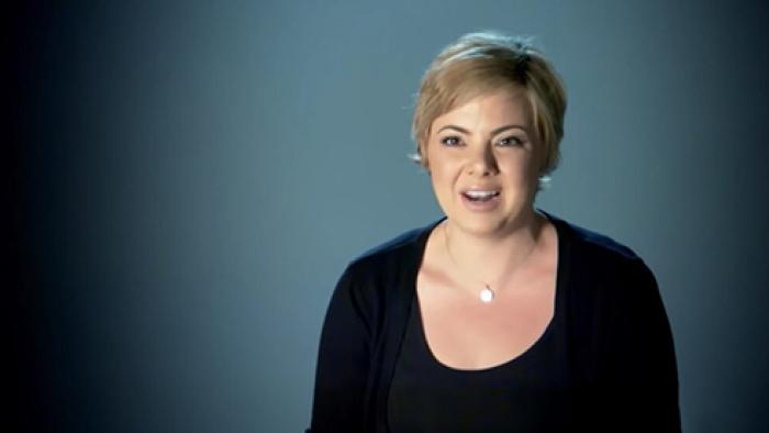 Scamanda TV show screenshot 3: Amanda Riley, in a black top and blonde pixie-cut, is pictured on a blue background