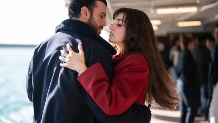 A brunette woman in a red coat embraces a dark-haired man in a black leather jacket in an airport-like setting