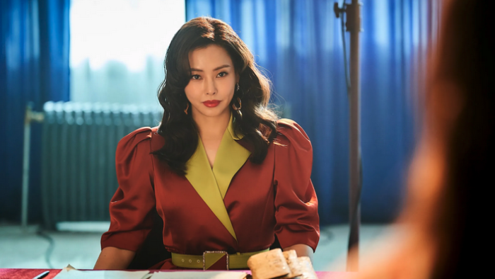 An Asian woman with curled hair in a leather blazer sits in what looks like a conference room and stars intensely at someone across the table