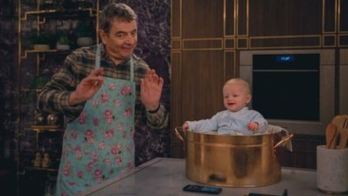 Man vs. Baby TV show sceenshot: A White man in a blue apron stands in front of a kitchen counter on which a baby in a onesie smiles in a metal pot