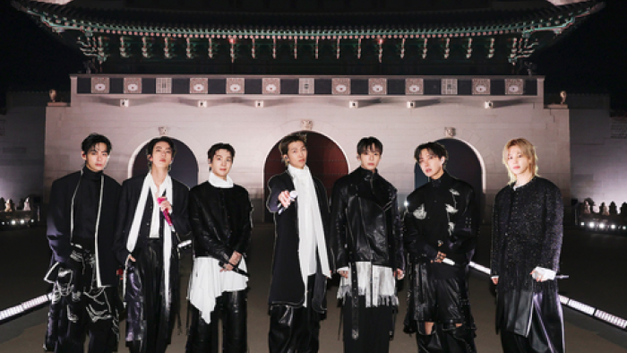 A group of performers stand side by side in coordinated dark outfits on a large stage, facing forward with confident posture, with an illuminated trad