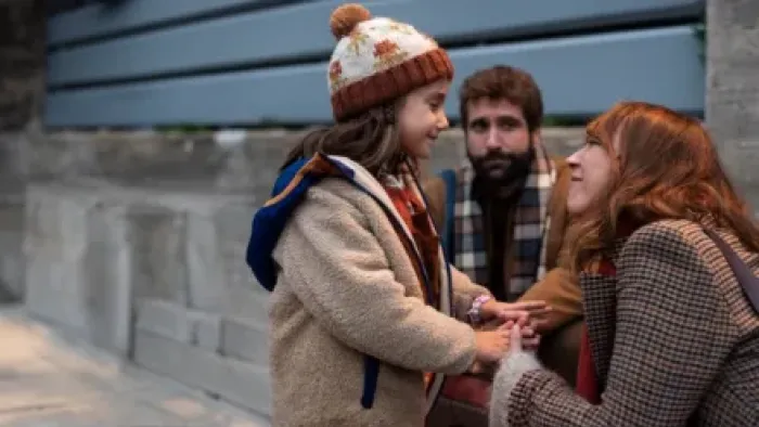 Shared Custody TV: Cris squats to Cloe's height and hold her hands, both smiling. Diego squats between them in the back, watching.